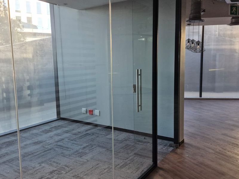 Modern open-plan office space featuring glass partitions, floor-to-ceiling windows, and a mix of carpet and laminate flooring with exposed industrial-style ceiling elements.