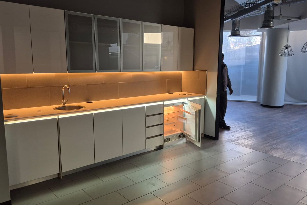 Modern office kitchenette featuring sleek white cabinetry, integrated under-counter refrigerator, stainless steel sink, and ambient under-cabinet lighting with industrial-style exposed ceiling ducting.