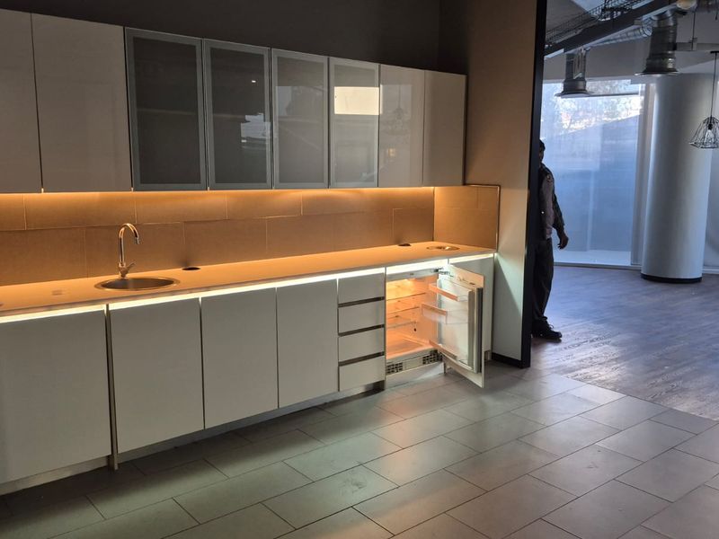 Modern office kitchenette featuring sleek white cabinetry, integrated under-counter refrigerator, stainless steel sink, and ambient under-cabinet lighting with industrial-style exposed ceiling ducting.