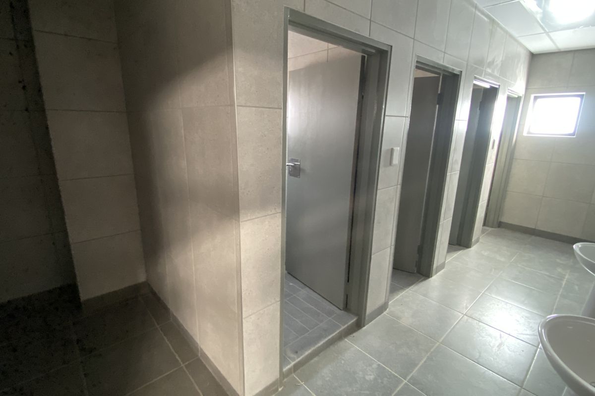 A clean, functional industrial-style communal bathroom featuring multiple toilet cubicles, tiled walls, and pedestal washbasins.