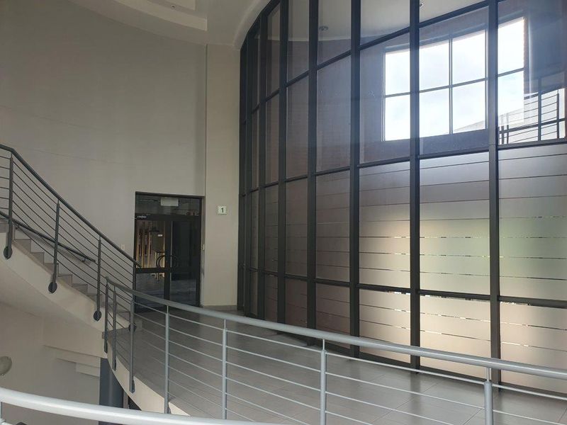 Spacious office building hallway featuring a striking curved glass curtain wall, modern metal staircase railing, and clean tiled flooring.