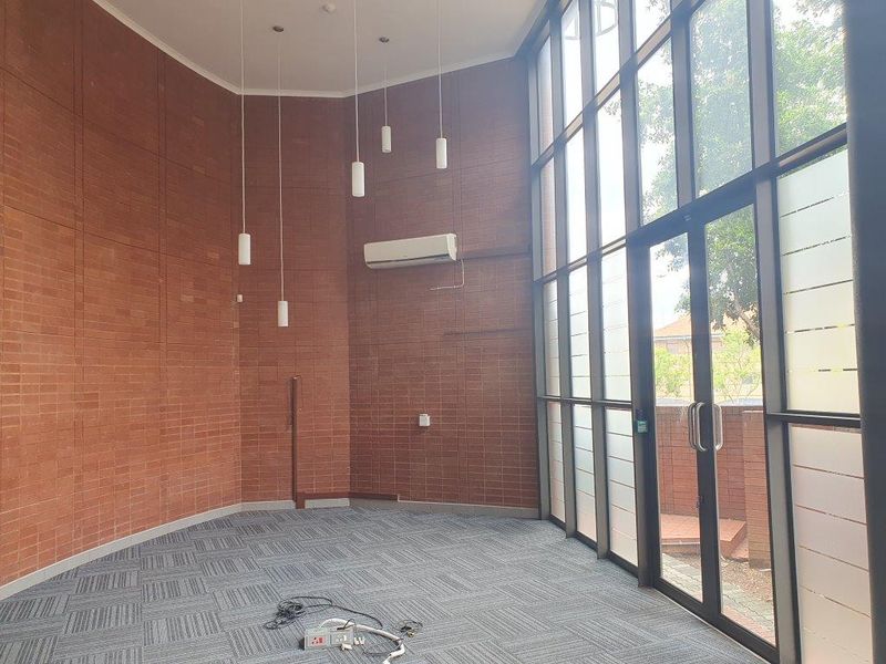 Bright, modern office space featuring exposed brick walls, high ceilings with pendant lighting, carpet tiles, and large floor-to-ceiling windows with glass door access.