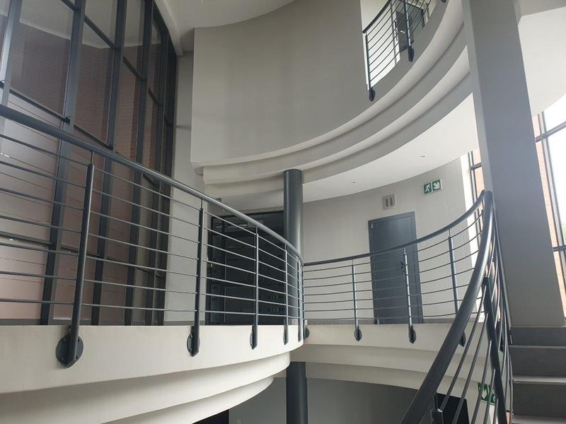 Modern office building interior featuring a curved staircase with metal balustrades, structural columns, and large glass partitions in a bright, open-plan lobby area.