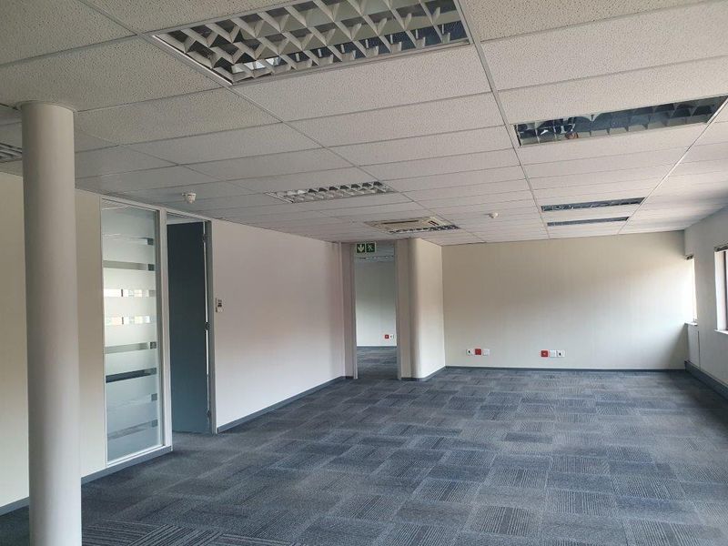 A spacious, empty open-plan office area featuring carpeted flooring, a suspended ceiling with recessed lighting, and a glass-paneled partition door.