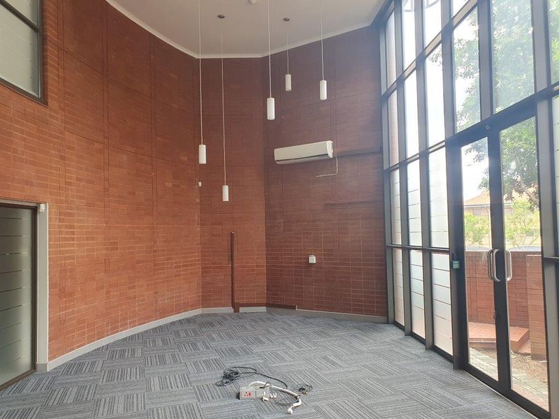 Spacious office area featuring exposed brick walls, high ceilings with pendant lighting, carpet tiles, and a large glass facade providing ample natural light.