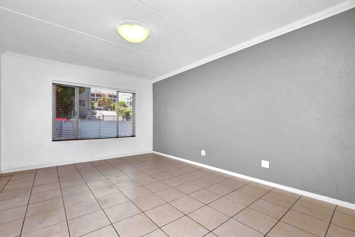 A bright, empty living room featuring neutral tiled flooring, a grey accent wall, and a large window with fitted blinds.