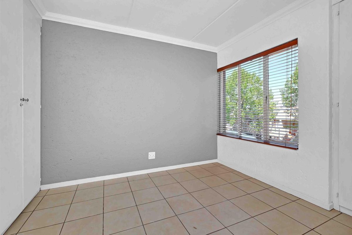 A bright, empty bedroom featuring neutral tiled flooring, a grey accent wall, and large windows fitted with horizontal blinds.