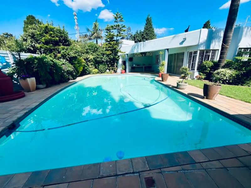 A well-maintained swimming pool area featuring a paved deck, lush garden surroundings, and a covered patio space ideal for outdoor relaxation.