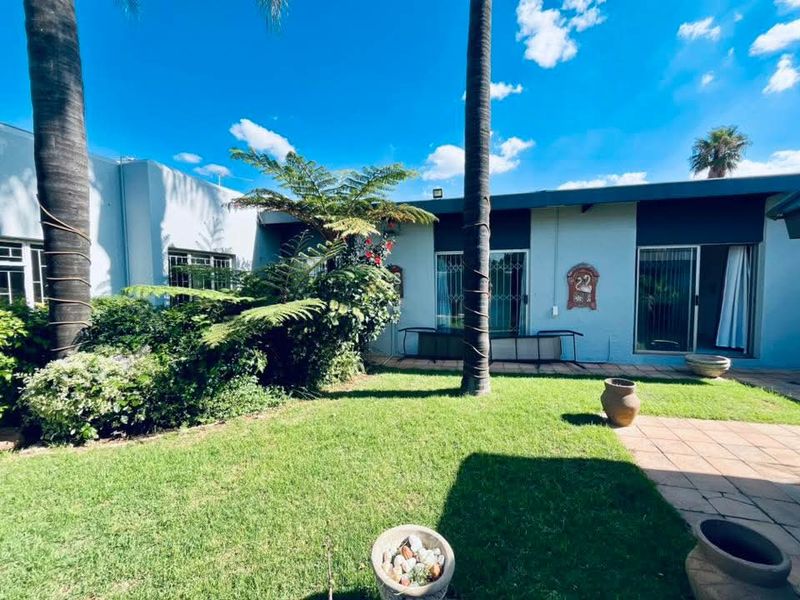 A rear garden view featuring a paved patio area, established lawn, mature palm trees, and sliding glass doors with security gates.