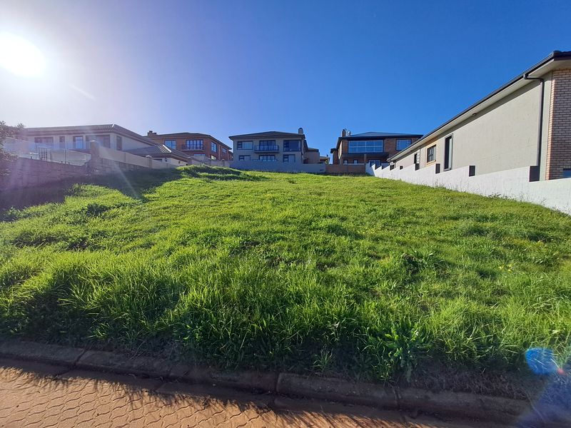 A vacant residential plot featuring a gentle upward slope, bordered by existing neighboring properties and a paved street curb.