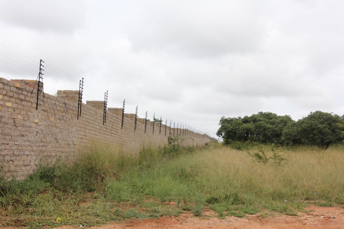 A vacant residential plot featuring a long perimeter boundary wall equipped with electric fencing and surrounding natural vegetation.