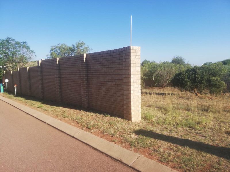 A view of the vacant residential plot featuring a sturdy brick boundary wall along the paved street frontage.