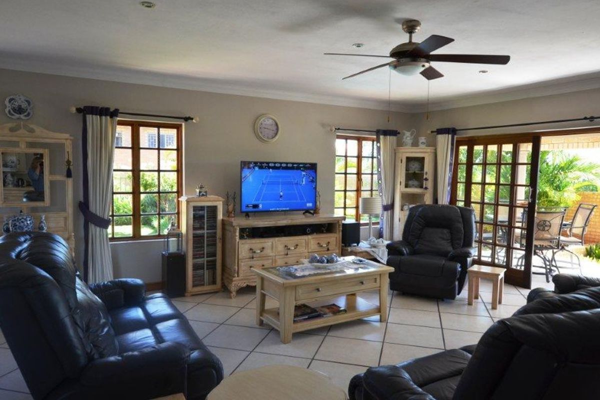 Spacious living room featuring tiled flooring, a ceiling fan, and large wooden-framed windows and sliding doors providing access to the outdoor patio area.