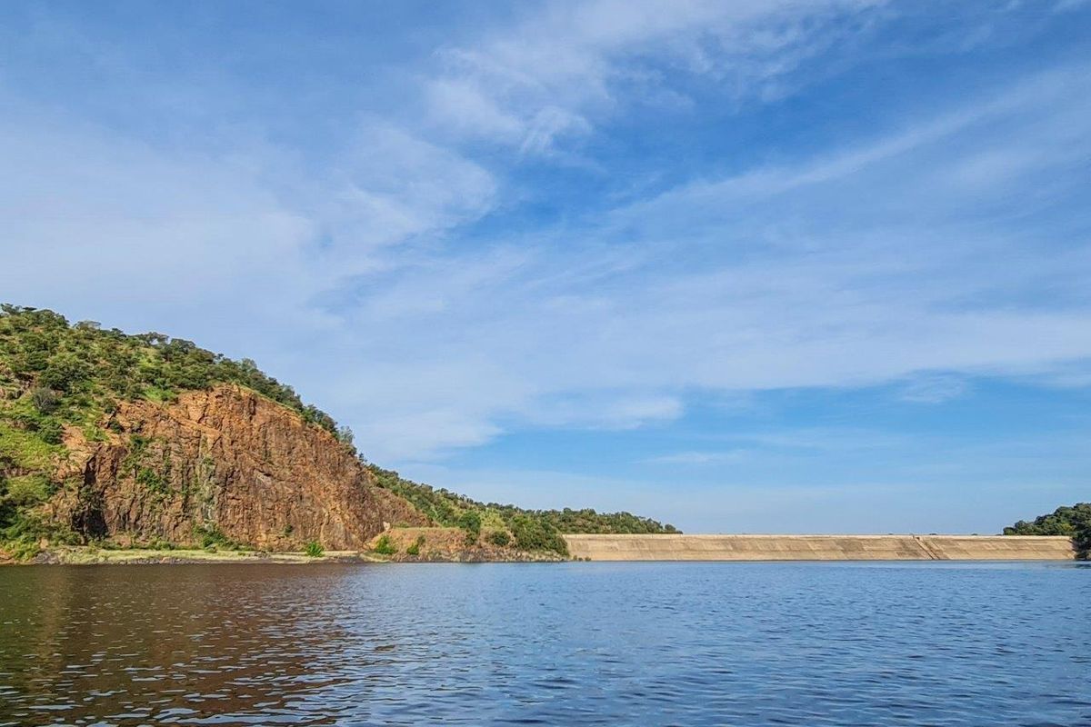 A scenic view of a large dam wall set against a natural rocky hillside and expansive water body.