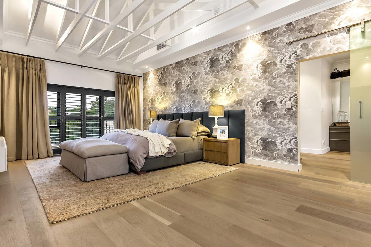 Spacious bedroom featuring vaulted ceilings with exposed beams, engineered timber flooring, a feature wallpaper wall, and a sliding glass door leading to a balcony.