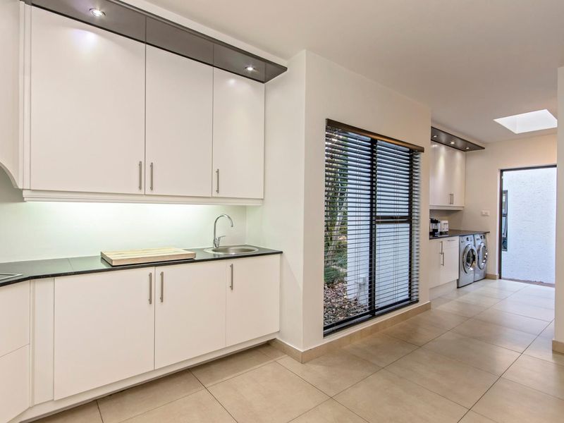 A modern, well-lit scullery featuring sleek white cabinetry, dark stone countertops, a stainless steel sink, and tiled flooring with a skylight.