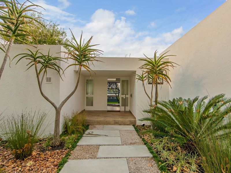 A modern, minimalist entrance featuring a paved walkway leading to a timber-decked porch and a glass-panelled front door.
