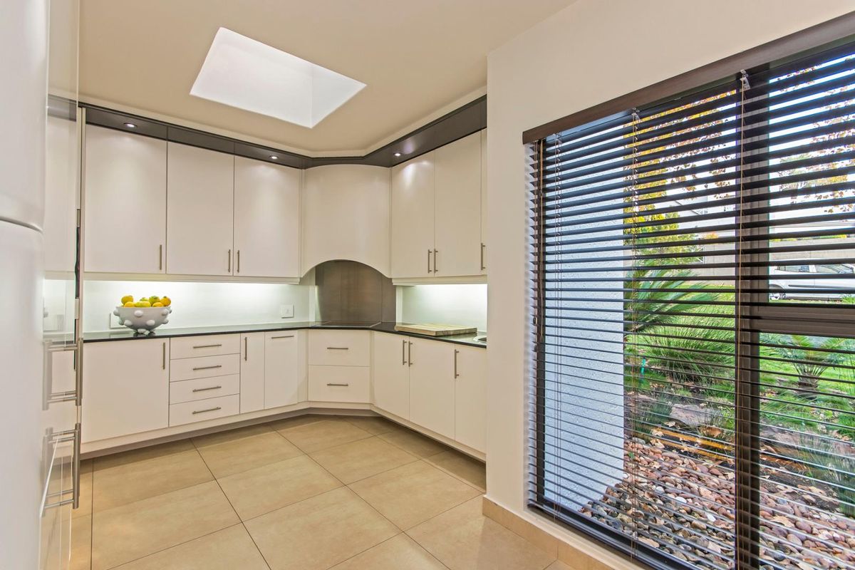 Modern kitchen featuring sleek white cabinetry, dark stone countertops, recessed lighting, a skylight, and large windows with venetian blinds overlooking the garden.