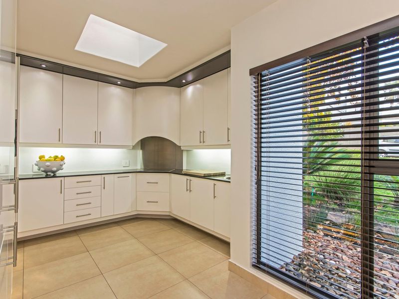 Modern kitchen featuring sleek white cabinetry, dark stone countertops, recessed lighting, a skylight, and large windows with venetian blinds overlooking the garden.