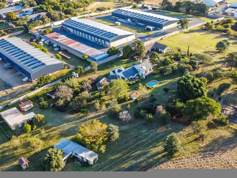 Aerial view of a farm property featuring a residential dwelling with a swimming pool, surrounded by extensive grounds and adjacent large-scale warehouse facilities.