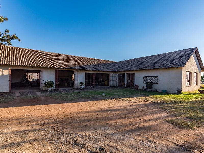 A wide-angle view of a farm outbuilding featuring multiple open garage bays, a tiled roof, and a large dirt and grass courtyard area.