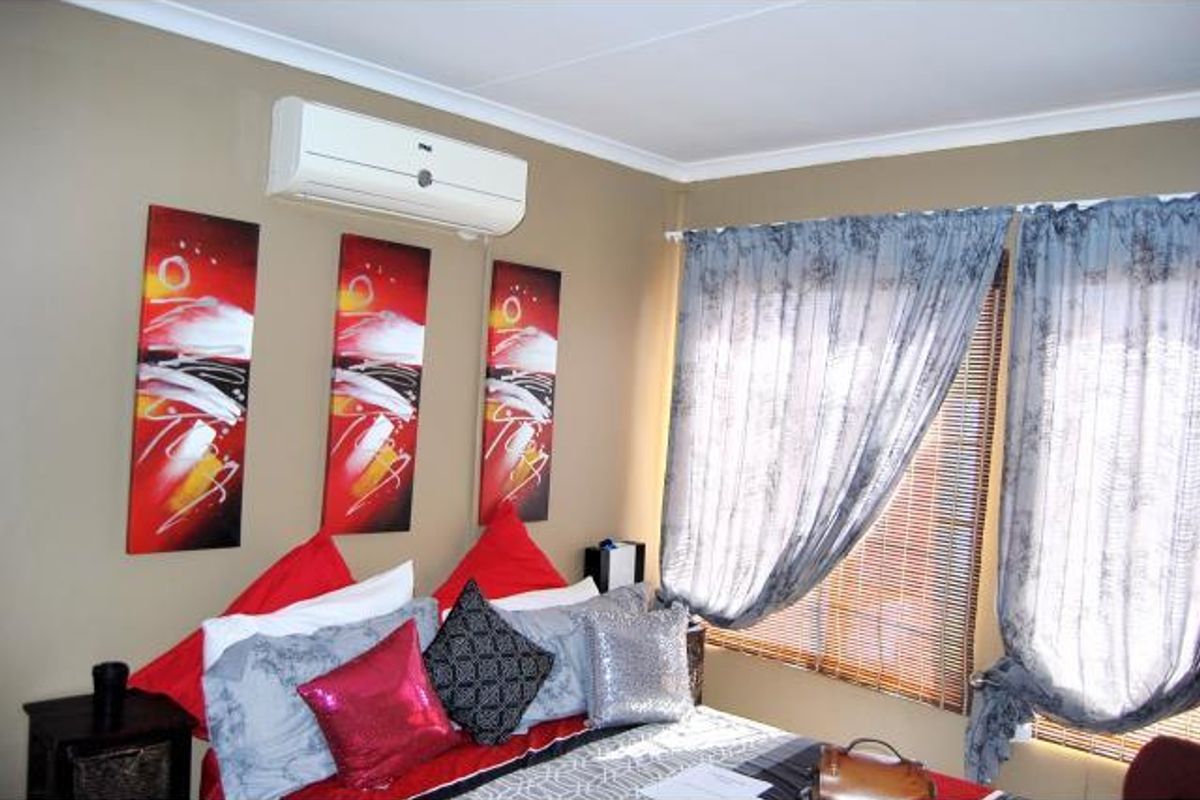 A bedroom featuring a wall-mounted air conditioning unit, large windows with fitted curtains and blinds, and neutral wall finishes.
