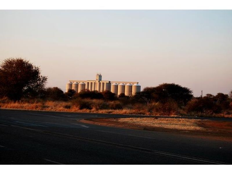 A view of industrial grain silos situated on a large commercial plot, visible from the adjacent asphalt road.