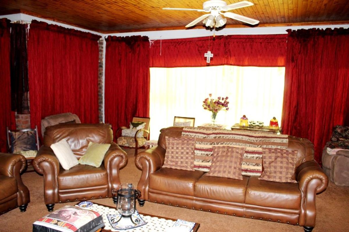 A living room featuring a timber-clad ceiling, ceiling fan, and large windows draped with heavy curtains.