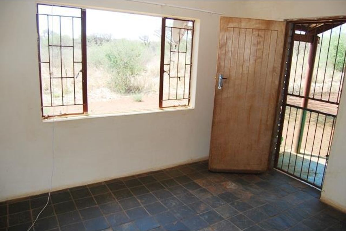 An interior view of a vacant room featuring dark tiled flooring, security bars on windows, and a wooden door leading to an outdoor area.