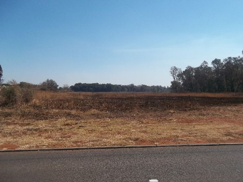A wide-angle view of a vacant residential plot featuring dry grass and natural vegetation, accessible from the adjacent paved road.