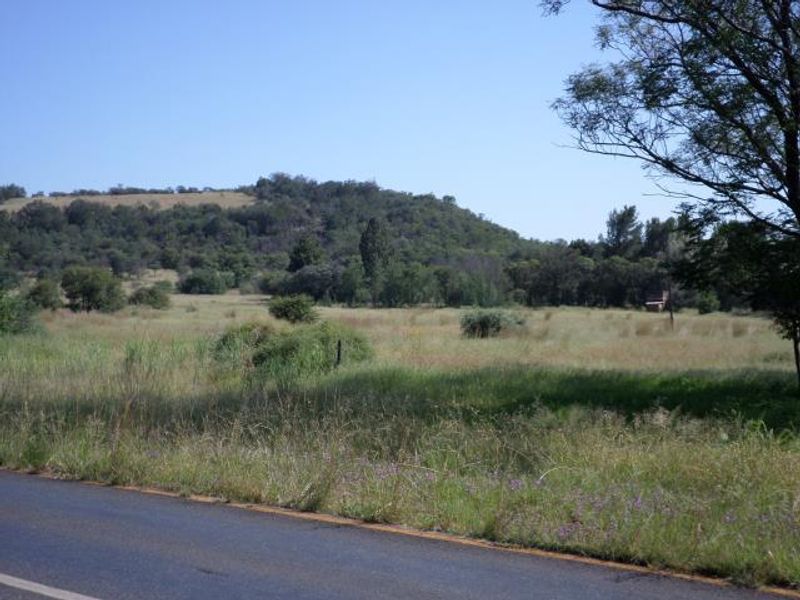 A view of a vacant residential land plot featuring natural grass, scattered trees, and a hilly landscape backdrop.