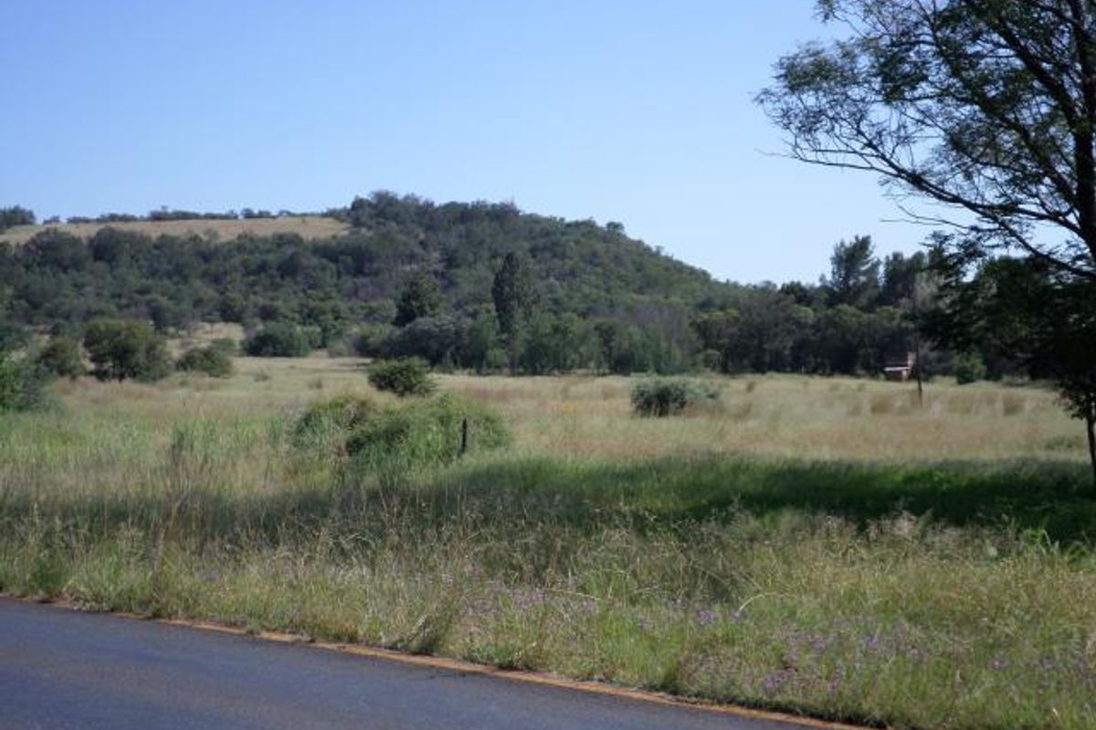 A view of a vacant residential land plot featuring natural grass, scattered trees, and a hilly landscape backdrop.