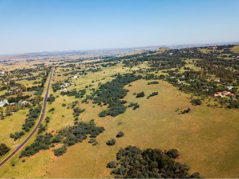 An aerial view of a large, undeveloped residential land parcel featuring rolling grassy terrain, scattered trees, and proximity to a main road.