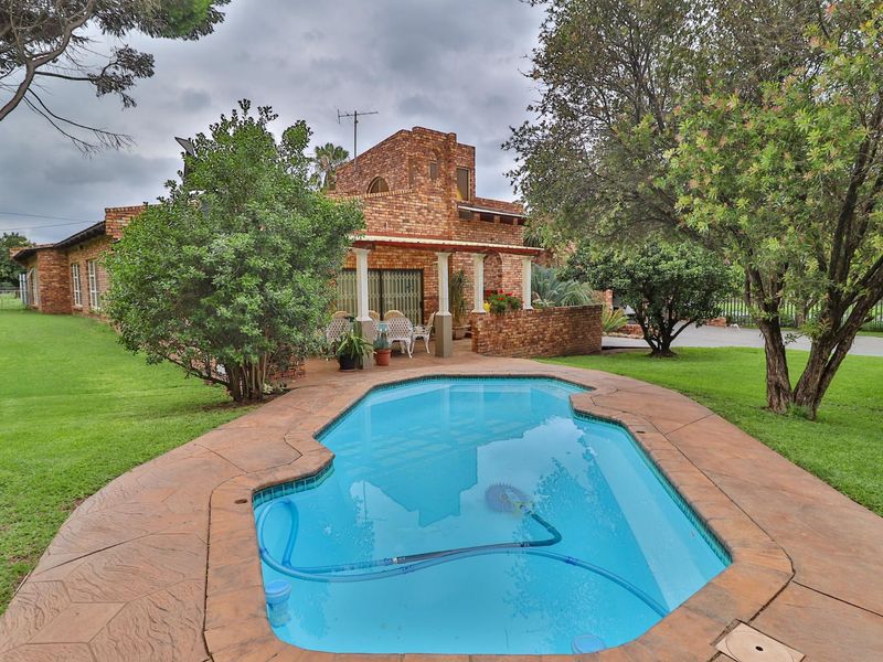 A well-maintained swimming pool area featuring a paved surround, established garden, and a covered patio attached to the face-brick dwelling.
