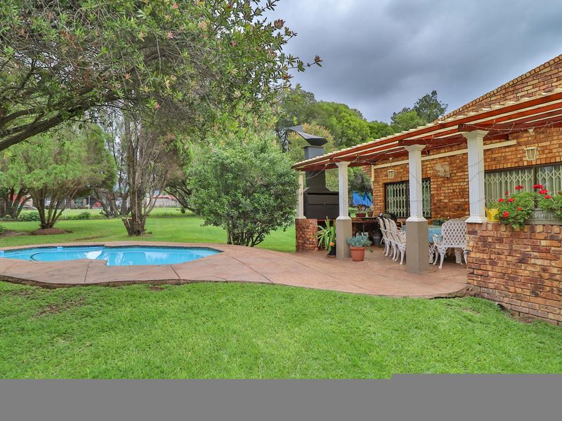 Covered brick patio featuring a built-in braai, overlooking a well-maintained garden and swimming pool area.