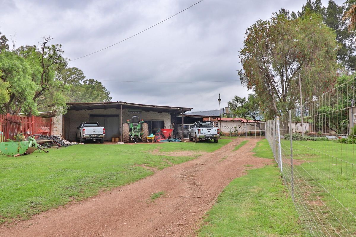 A view of the rear exterior area of the small holding, featuring a large open-sided metal structure, dirt driveway, and wire mesh perimeter fencing.