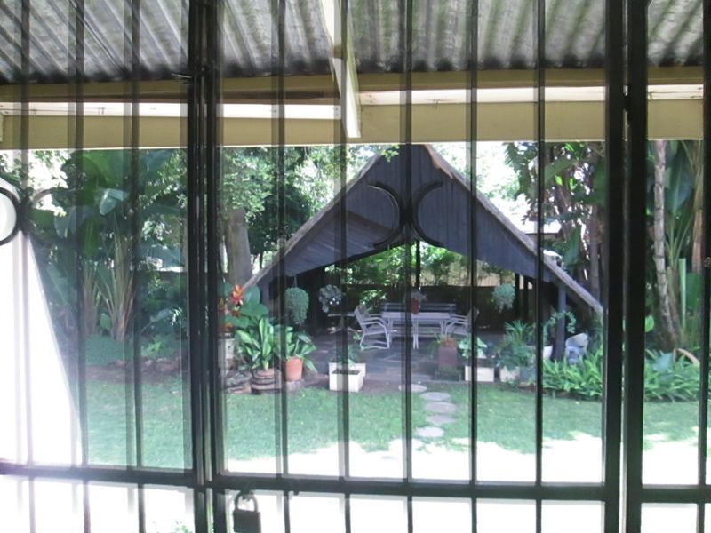 View from behind a security gate looking out onto a lush garden featuring a covered outdoor entertainment structure and paved pathway.
