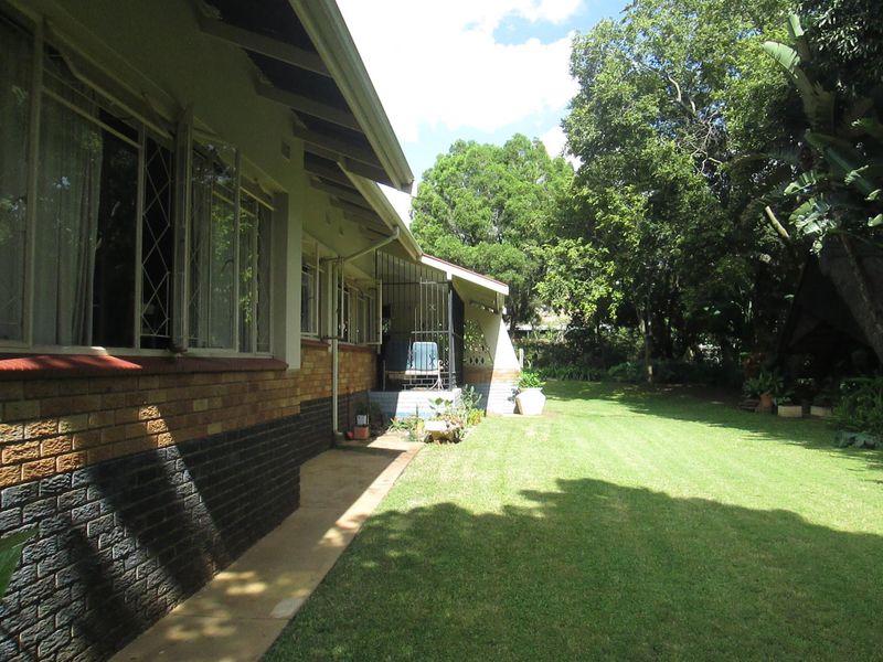 A view of the rear garden featuring a well-maintained lawn, mature trees, and a covered patio area attached to the brick house.