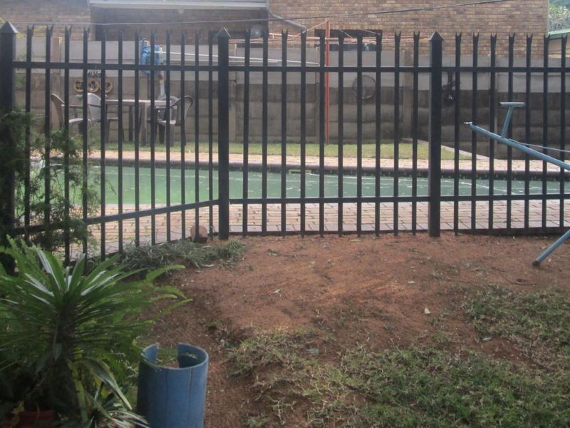 Outdoor pool area featuring a swimming pool with paved surround, enclosed by black metal security fencing and a brick boundary wall.