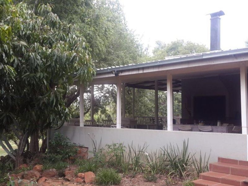A covered outdoor patio area featuring a built-in braai and tiled steps, overlooking the surrounding garden and mature trees.