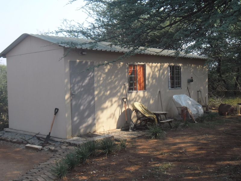 A simple detached outbuilding or cottage featuring a corrugated metal roof, security-barred windows, and a paved walkway set in a natural garden environment.