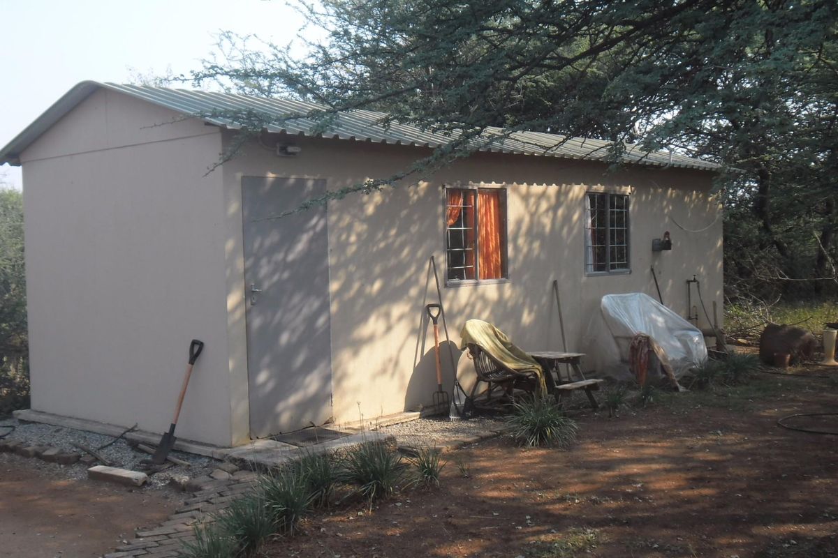 A simple detached outbuilding or cottage featuring a corrugated metal roof, security-barred windows, and a paved walkway set in a natural garden environment.