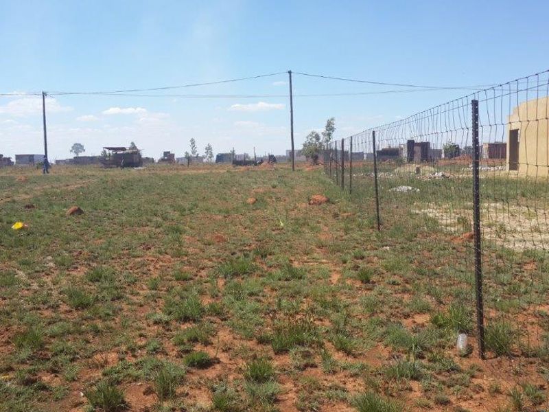 A view of a vacant residential plot featuring wire mesh perimeter fencing and overhead power lines, set within a developing neighborhood.