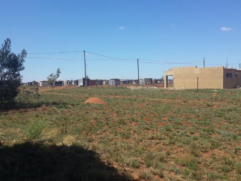 A vacant residential plot featuring natural terrain, perimeter fencing, utility connections, and an unfinished masonry structure on site.