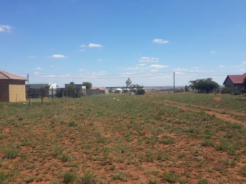 A vacant residential plot featuring natural grass cover, perimeter wire fencing, and visible overhead power lines in a suburban setting.