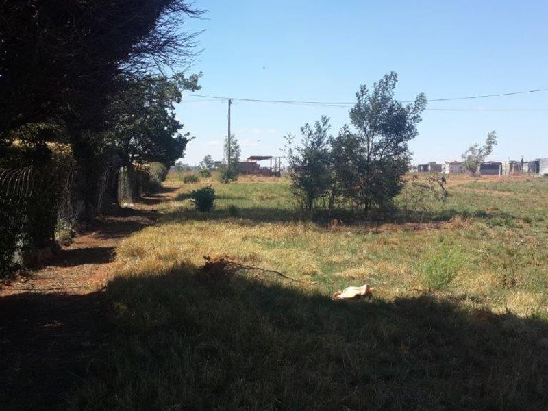 A view of a vacant residential plot featuring natural grass, scattered trees, and perimeter wire fencing under a clear sky.