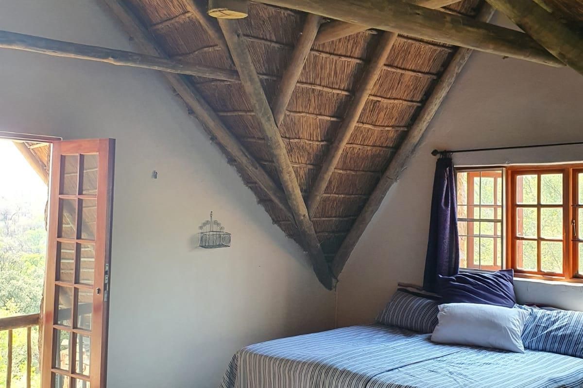 A rustic bedroom featuring a high vaulted thatched roof with exposed timber beams, wooden flooring, and direct access to an outdoor balcony.
