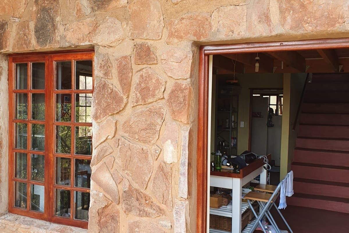 Exterior view of a rustic stone-clad house featuring a timber-framed window and an open doorway leading into an interior living space with a staircase.