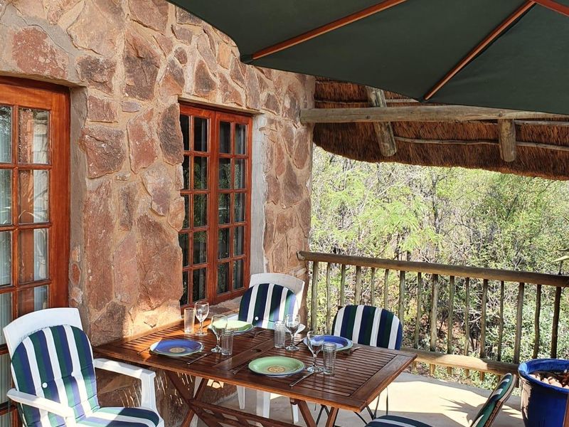 A covered outdoor patio featuring rustic stone wall cladding, a traditional thatch roof, and a timber balustrade overlooking the surrounding natural landscape.