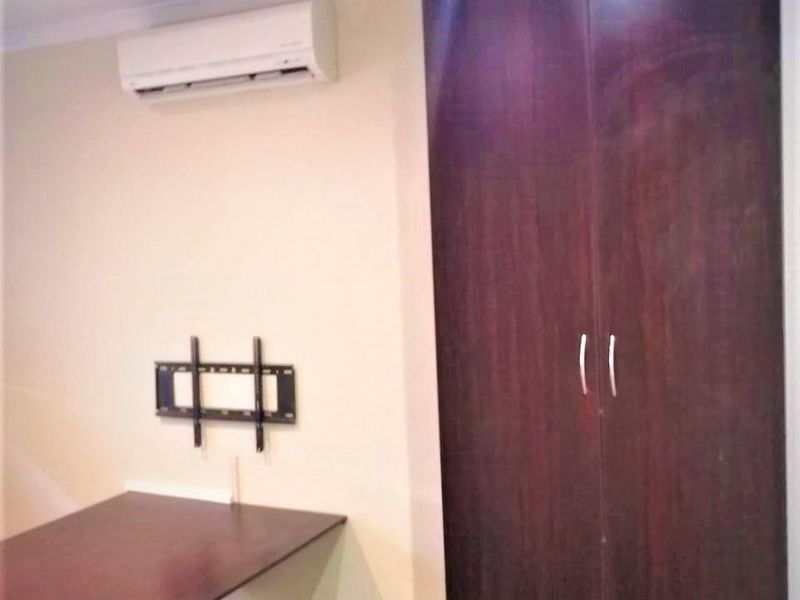 A functional bedroom space featuring a built-in dark wood wardrobe, wall-mounted air conditioning unit, and a fixed desk surface.